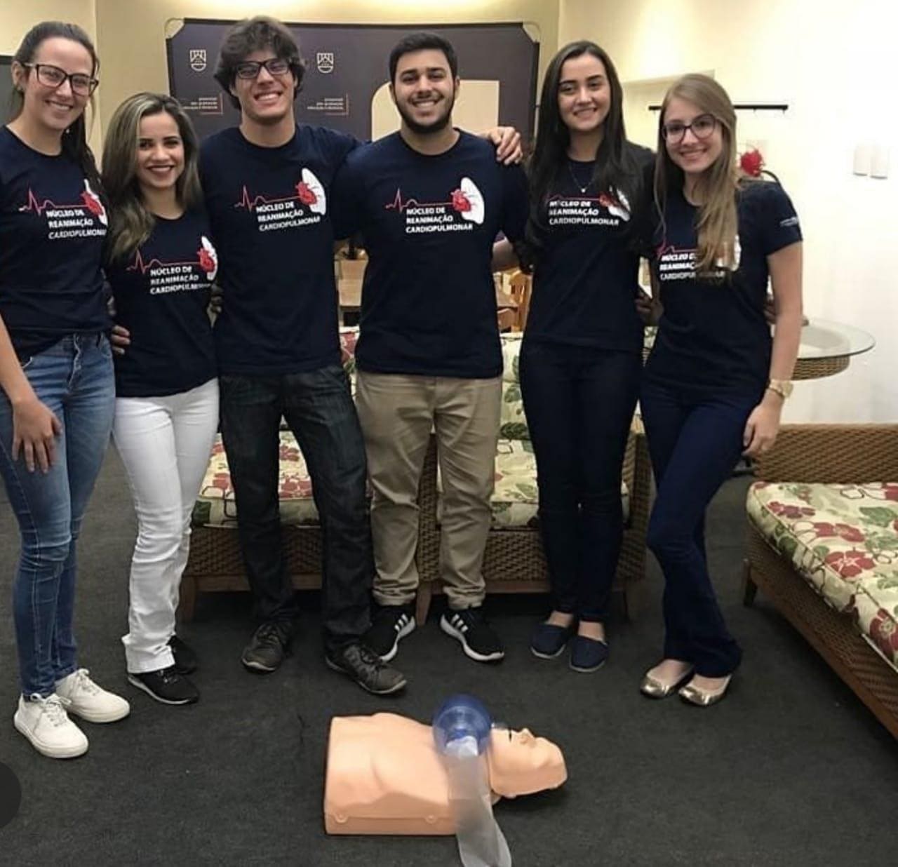 Imagem representando 6 pessoas e um boneco para testes de reanimação cardiopulmonar tirando foto